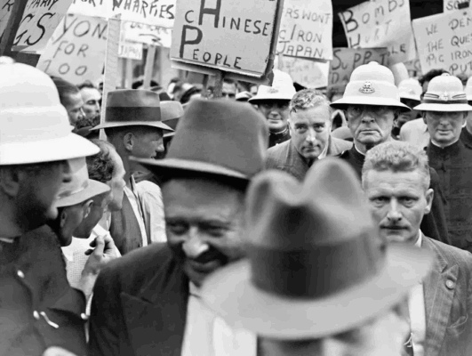 In 1939 photo, Attorney-General Menzies walks through strikers. Posters include: "Bombs Hurt People". 