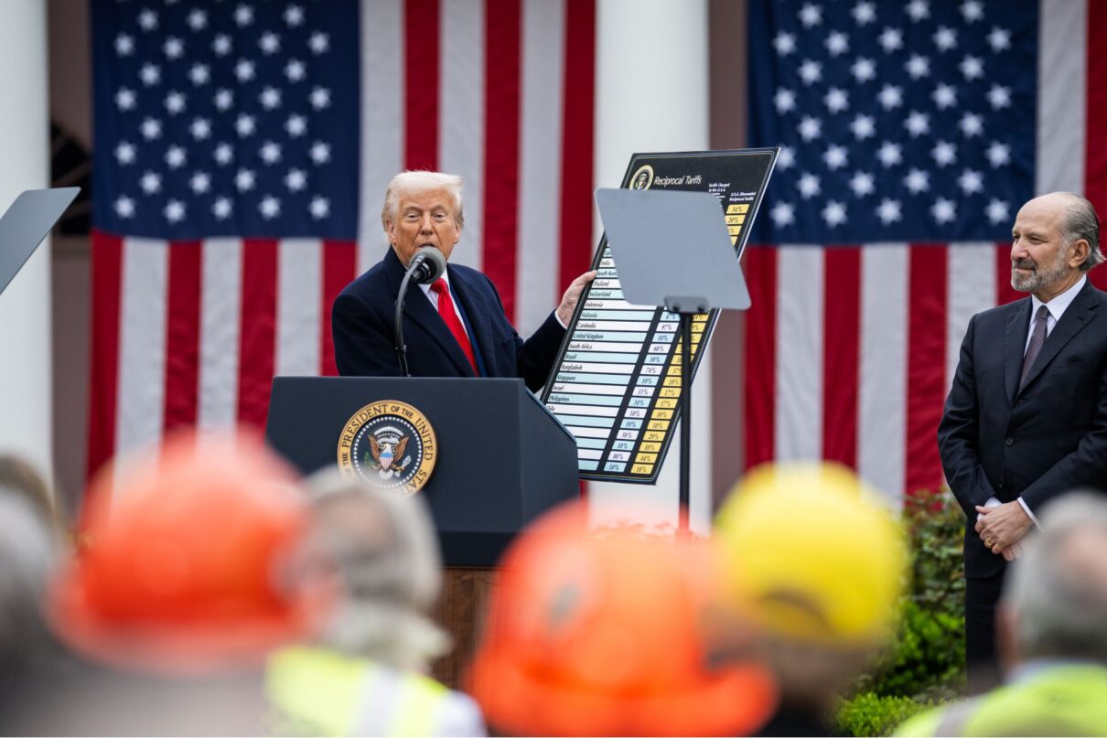 Donald Trump holding a sign with tariff levels on a podium in front of large American flags